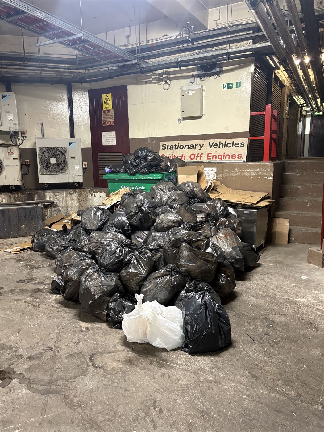 Commercial bin store before clearance, waste bags heaped beside the general waste bin